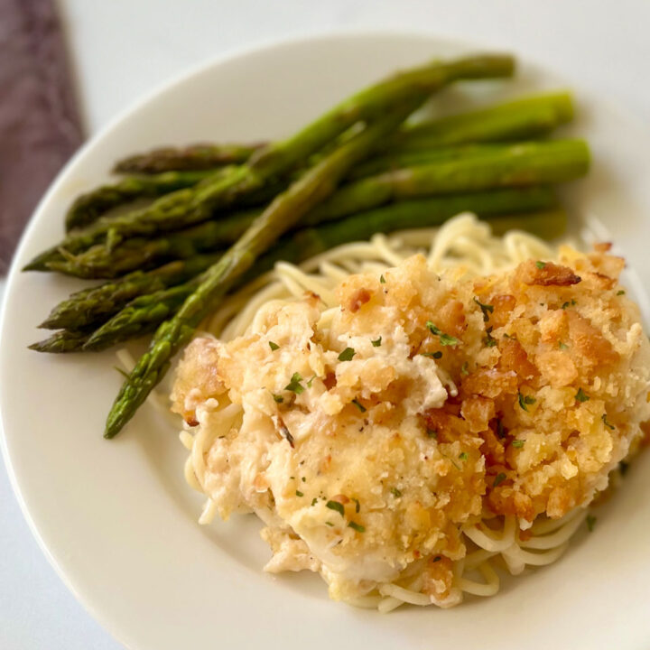 There is a white plate with baked scallops with a cracker crumb topping laying on a bed of angel hair pasta. There are stalks of asparagus on the plate. There is a gray cloth napkin in the background. Items are on a white marbled surface.