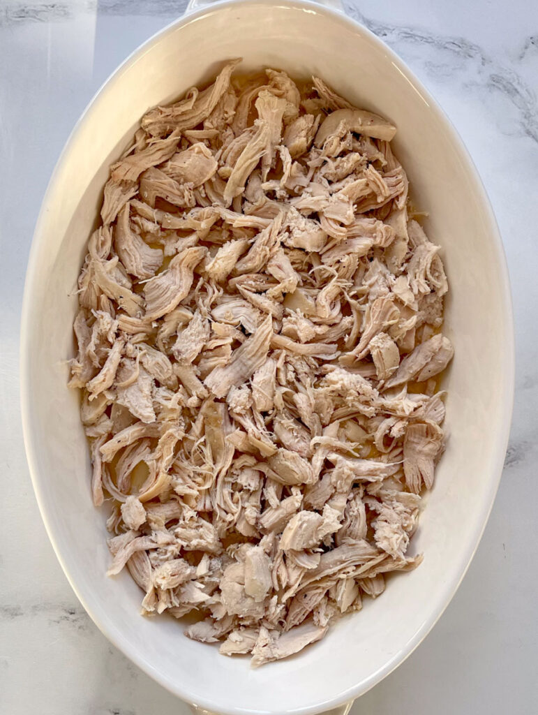 There is a large white oval dish with a layer of shredded chicken covering a layer of melted butter. The dish is on a white marbled surface.
