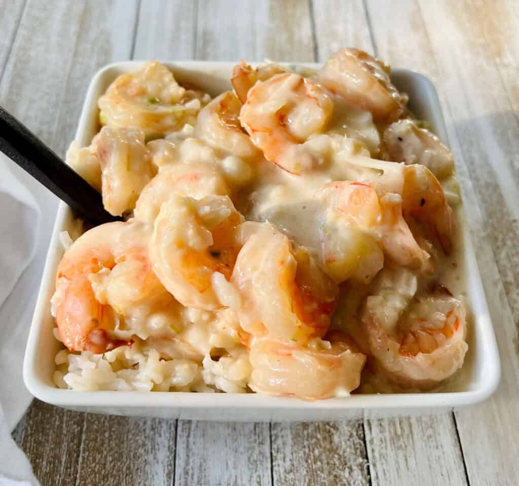 There is a square white dish with white rice, cooked shrimp in a white cream sauce. There is a spoon resting in the dish. The dish is on a wood surface. There is a white cloth napkin laying next to the dish.