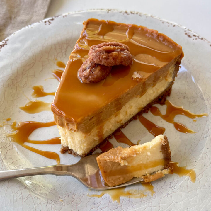 There is a slice of cheesecake that has a light brown creamy top layer and a dark brown cookie crust on a white plate. There is caramel sauce drizzled over the top of the cheesecake. There are 2 sugar coated pecans sitting on top. There is a fork with some of the cheesecake on it, laying on the plate.