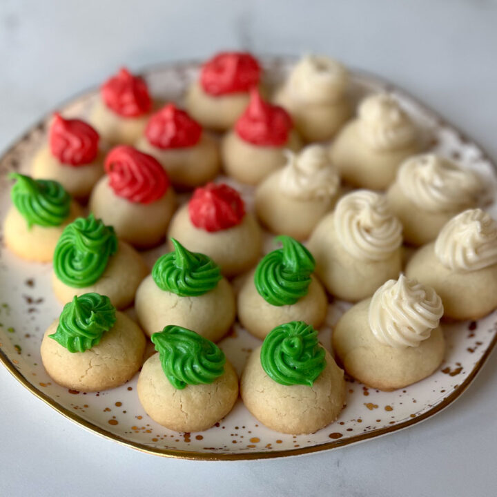 There are twenty one round white cookies on a round white plate that has a gold rim and tiny gold specks on it. Six of the cookies have a small swirl of green frosting on them. Seven of the cookies have a small swirl of red frosting on them. Seven of the cookies have a swirl of white frosting on them. The plate is on a white marbled surface.