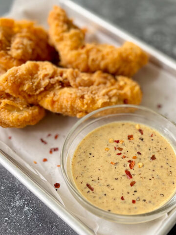 There is a small clear glass bowl with a yellow creamy sauce in it. There are small red pepper flakes sprinkled on top of the sauce. The bowl is on a stainless steal rectangular tray lined with white paper. There are a few pieces of fried chicken strips on the tray. The tray is on a dark gray surface.