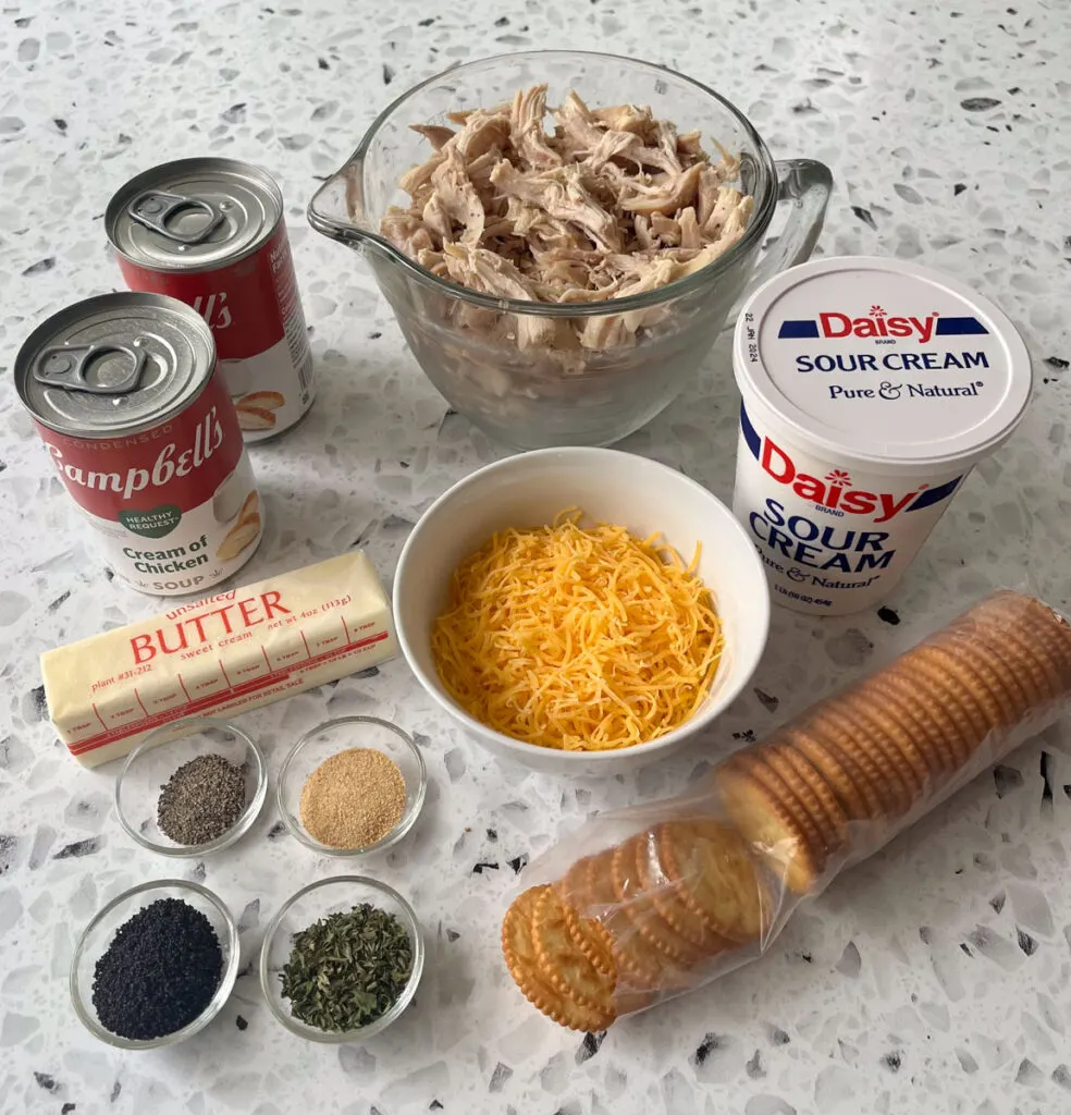 There is a large measuring bowl with shredded cooked chicken. There are two cans of condensed soup, a white carton of sour cream, a stick of butter, a white bowl with yellow shredded cheese in it, four very small clear glass bowls with seasonings in them, and a clear plastic sleeve of light brown round crackers. Items are on a white and black terrazzo surface.