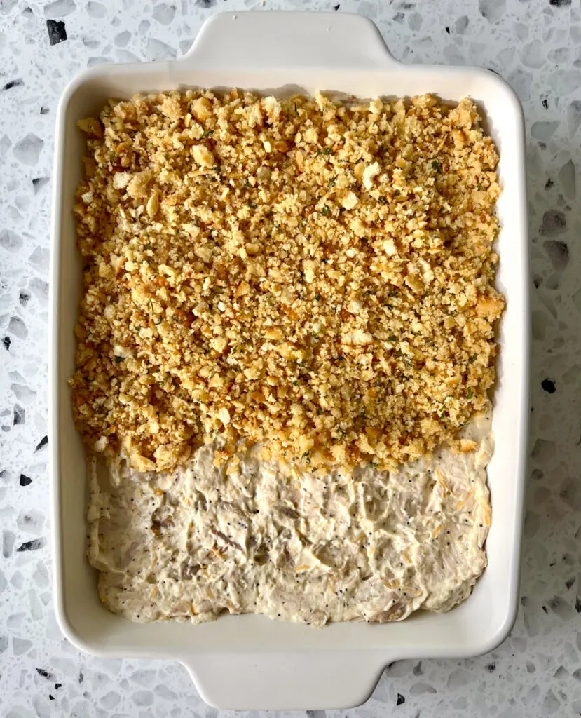 There is a white rectangular baking dish with a creamy mixture made up condensed soup, sour cream, shredded cheese, shredded chicken and seasonings in it. There is a cracker crumb topping on half of the chicken mixture. The baking dish is on a white and black terrazzo surface.