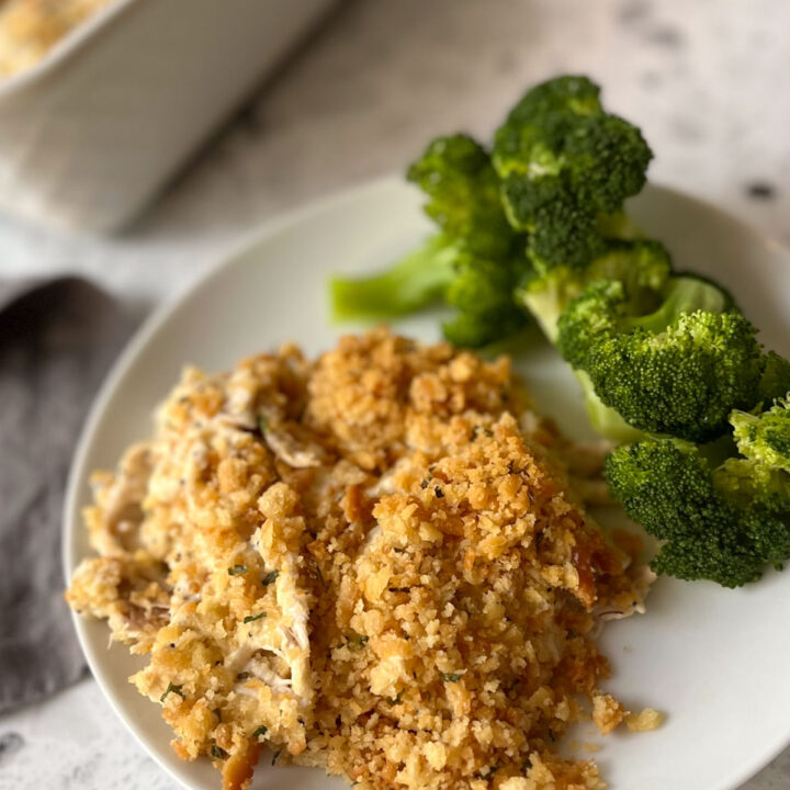 There is a white plate with a creamy chicken mixture and pieces of green broccoli on it. There is part of a white rectangular baking dish with a creamy white chicken mixture with a cracker crumb topping in it. There is a stainless steel spoon resting in the baking dish. There is a dark gray cloth napkin laying next to the plate. Items are on a white and black terrazzo surface.