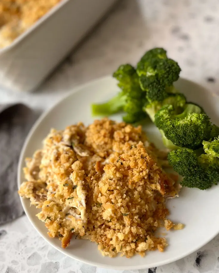 There is a white plate with a creamy chicken mixture and pieces of green broccoli on it. There is part of a white rectangular baking dish with a creamy white chicken mixture with a cracker crumb topping in it. There is a stainless steel spoon resting in the baking dish. There is a dark gray cloth napkin laying next to the plate. Items are on a white and black terrazzo surface.