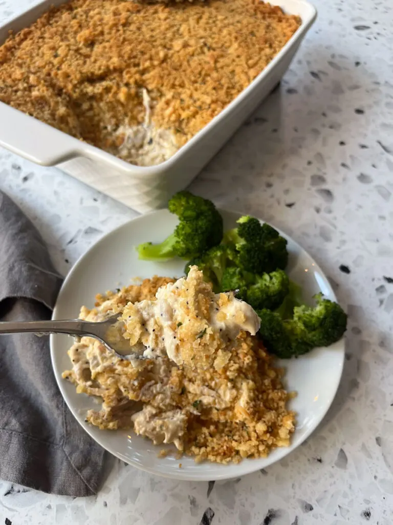 There is a white plate with a creamy chicken mixture and pieces of green broccoli on it. There is a fork resting on the plate. There is a white rectangular baking dish with a creamy white chicken mixture with a cracker crumb topping in it. There is a dark gray cloth napkin laying next to the plate. Items are on a white and black terrazzo surface.