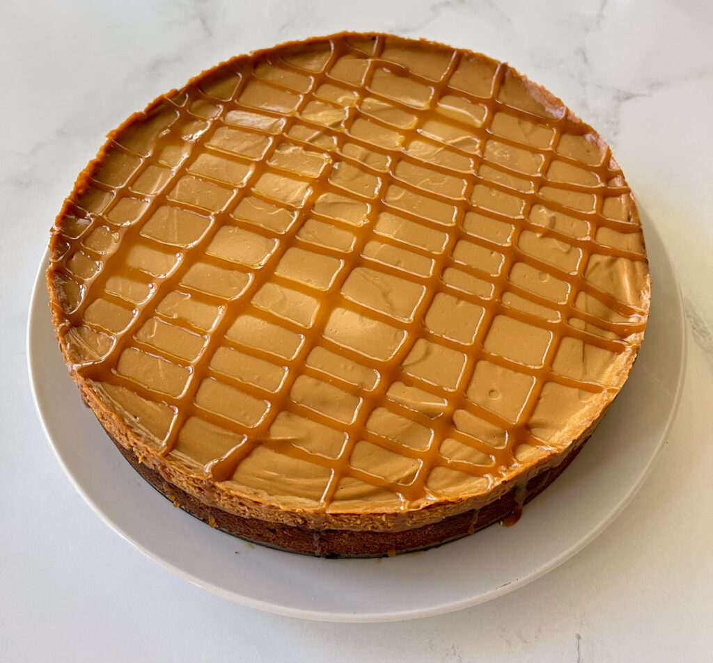 There is a round cheesecake on a large round white platter. The cheesecake has a dark brown cookie crust and a light brown creamy top layer. There is caramel sauce drizzled over the top. The platter is on a white marbled surface.