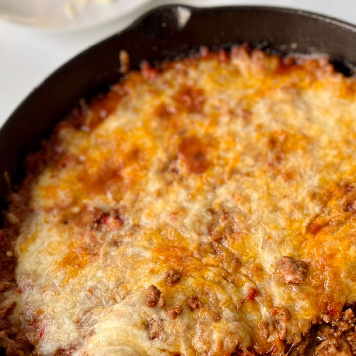 There is a black cast iron skillet with a meat mixture with melted cheese on top. There is a stainless steel spoon resting in the skillet. There is a white plate in the background. Items are on a white marbled surface.