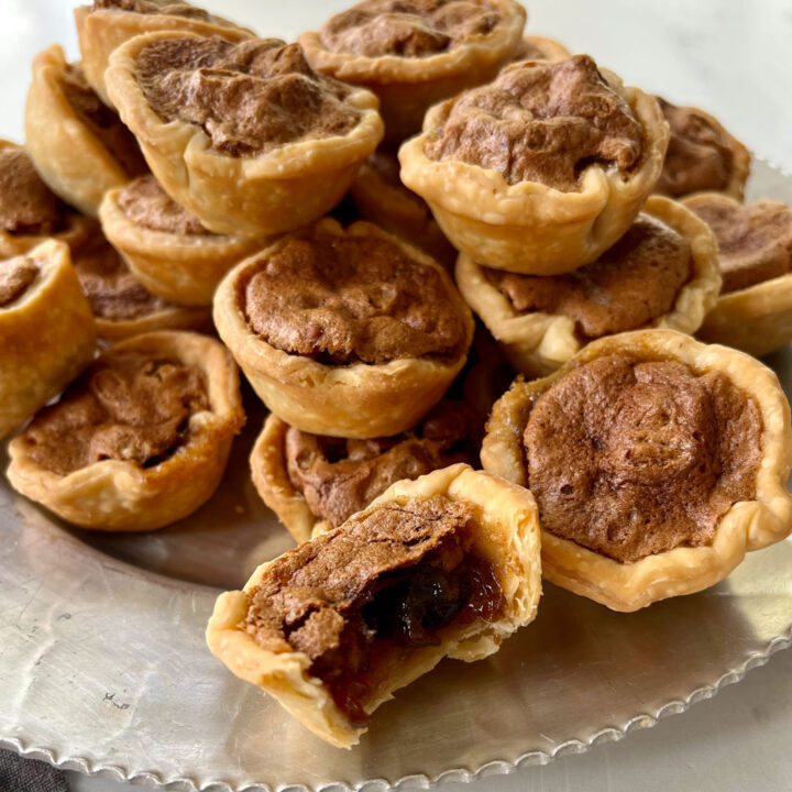There is a sliver plate filled with baked mini pie shaped shells with brown sugar, pecan pieces, and brown sugar in them. The plate is on a white marbled surface.