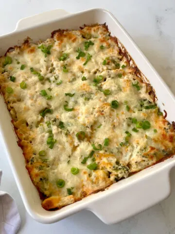There is a white rectangular dish with a baked shredded chicken, broccoli, cauliflower, and creamy cheese mixture in it. There are chopped small pieces of green onion on the top of the mixture. The dish is on a white marbled surface.