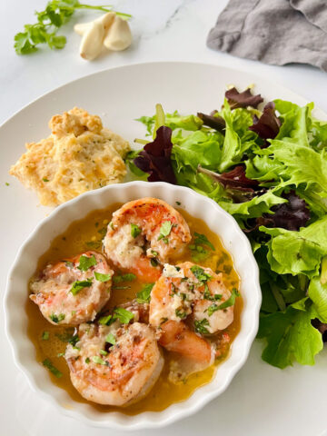 There is a white plate with a small shallow bowl with four large cooked shrimp resting in a pool of light yellow sauce. There is grated white cheese and chopped green parsley on top of the shrimp. There is a pile of leafy salad greens and a biscuit on the plate. There are three pieces of garlic cloves, a sprig of green leafy parsley, and a dark gray cloth napkin in the background. Items are on a white marbled surface.