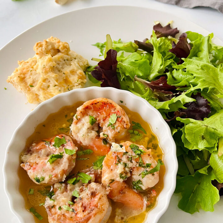 There is a white plate with a small shallow bowl with four large cooked shrimp resting in a pool of light yellow sauce. There is grated white cheese and chopped green parsley on top of the shrimp. There is a pile of leafy salad greens and a biscuit on the plate. There are three pieces of garlic cloves, a sprig of green leafy parsley, and a dark gray cloth napkin in the background. Items are on a white marbled surface.