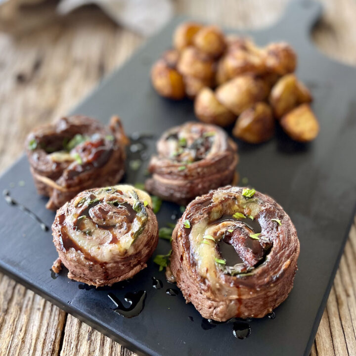 There is a black board with four 2-inch thick pieces of cooked steak rolled around white cheese and spinach. There is finely chopped green parsley sprinkled over the steak rolls. There is also a dark brown sauce drizzled over the steak rolls. There is a pile of cut up cooked potatoes behind the steak rolls. The black board is on a wood surface.