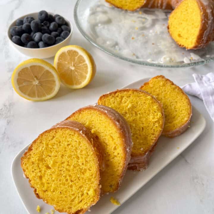 There is a small rectangular tray with slices of yellow cake on it. In the background, there is a large clear glass plate with half of a round, ring-shaped cake with grooved sides. There is a white glaze drizzled over the cake. There is also a small white bowl with blue berries and a lemon cut in half in the backgound. Items are on a white marbled surface.