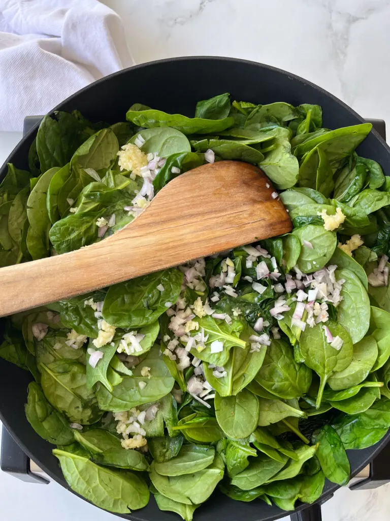 There is a large dark gray pan with spinach, small pieces of onion and garlic cooking. There is a wooden spoon resting in the pan.