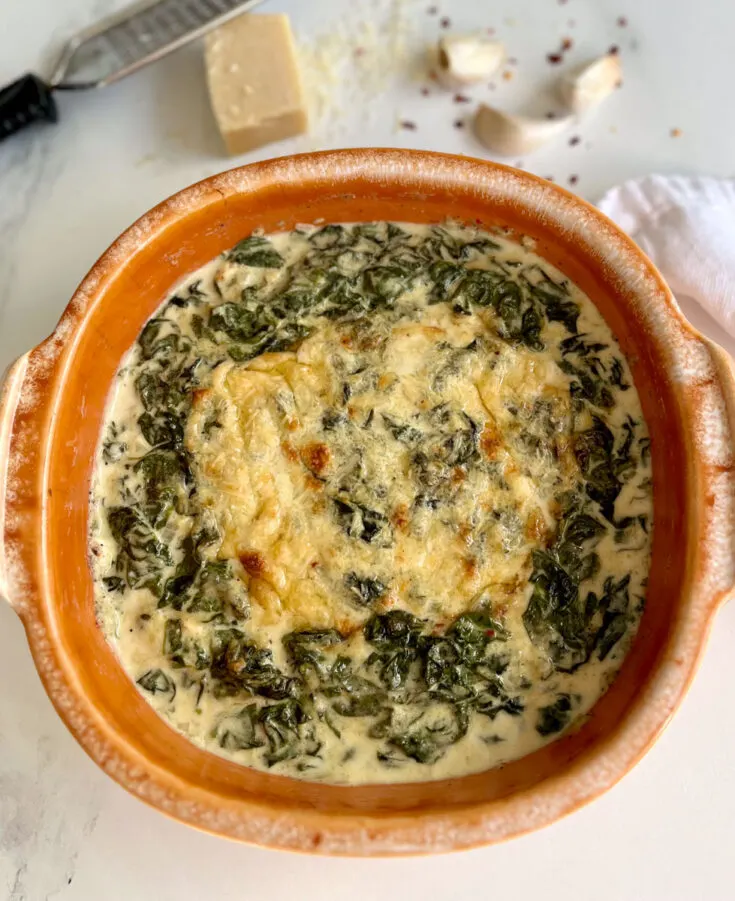 There is an oval orange baking dish with a baked creamy cheese spinach mixture in it. There are 3 cloves of garlic, a partial block of white cheese with a small flat grater, and red pepper flakes laying behind the dish. Items are on a white marble surface.