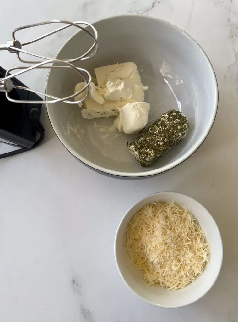 There is a large white mixing bowl with white cream cheese and a block of white cheese with green herbs in it. There is a hand mixer with metal beaters next to the bowl. There is a small bowl with shredded light yellow cheese in it sitting in front of the large mixing bowl. Items are on a white marbled surface.