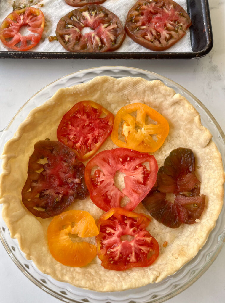 There is a clear glass pie pan with a baked pie crust in it. There is a layer of sliced tomatoes on the bottom of the crust. There is a baking pan lined with white paper towels with a layer of slice tomatoes on it in the background. Items are on a white marbled surface.