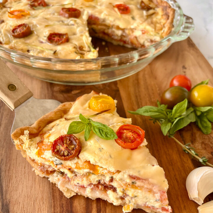 There is a slice of a piece of tomato pie which has layers of tomato and and a cheese mixture. The piece of pie is on a metal pie spatula. In the background there is a clear glass pie pan with sliced tomatoes and a cheese mixture in it topped with thin slices of light yellow cheese, slices of small tomatoes, and thin slices of green onions on top with a wedge cut out of it. Items are on a wood surface.