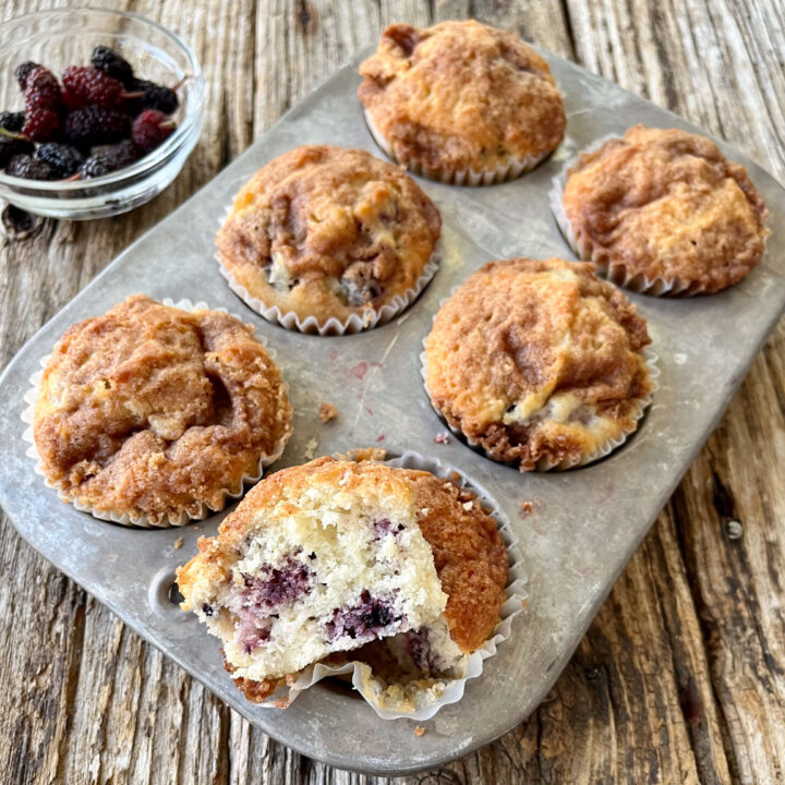 There is a metal 6 cup muffin tin with baked muffins in white cup liners in the muffin cups. One of the muffins is cut in half to expose the center of the muffin. There is a small glass bowl with red oblong berries in it in the background. Items are on a wood surface.