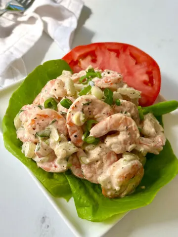 There is a square white plate. On the plate there is a scoop of cooked shrimp mixed with a creamy sauce. There are thin slices of green onion on top. The shrimp are sitting on a bed of green lettuce. There are slices of red tomato also on the plate. There is a white cloth napkin and a silver fork in the background. Items are on a white marbled surface.