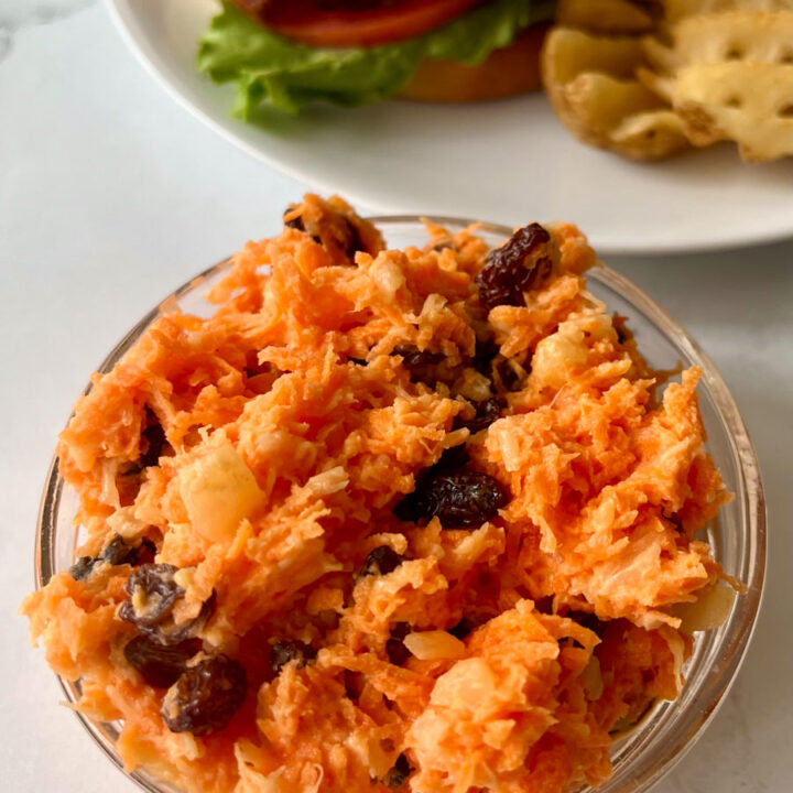 There is a clear glass bowl with carrot raisin salad in it. In the background there is a white plate with a a fried chicken sandwich and french fries on it. Items are on a white surface.