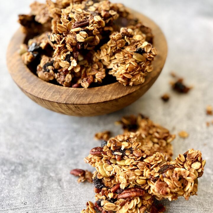 There is a wooden bowl with granola in it. The bowl is on a light gray surface. There are pieces of granola laying around the bowl.