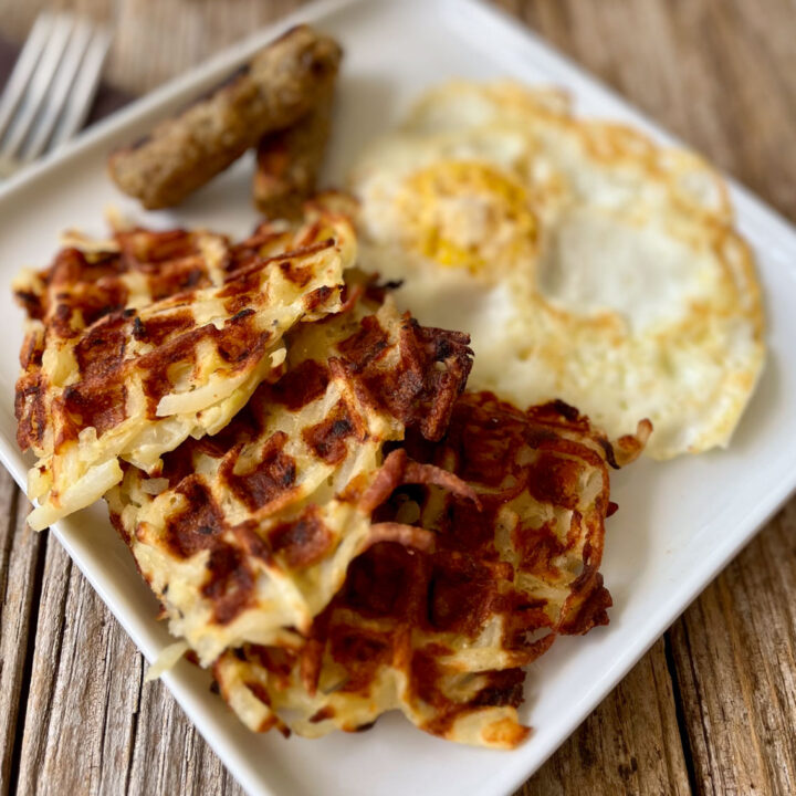 White square plate with hash brown patties, sausage links, and a fried egg on it. Plate is on a wood surface with a fork laying next to the plate.