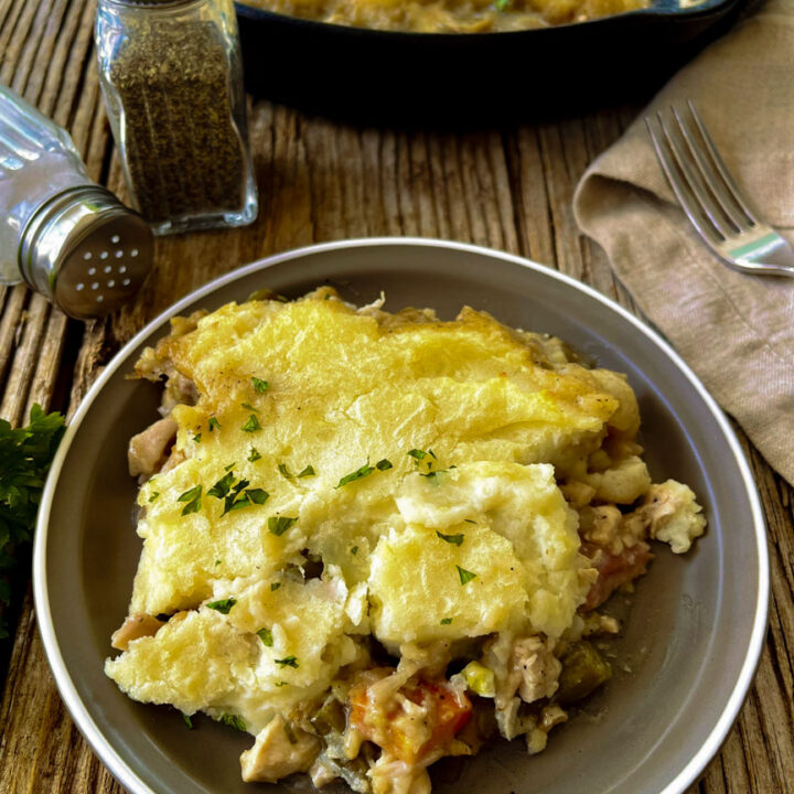 There is a brown plate with a serving of chicken with mashed potatoes pie on in. In the background there is a set of salt and pepper shakers, a fork on a brown cloth napkin and a large cast iron skillet with chicken mashed potatoes pie in it. Items are on a wood surface.