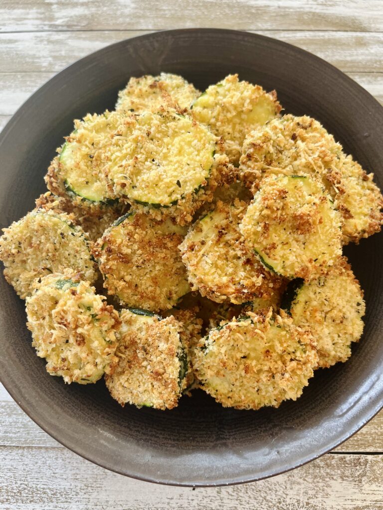 There is a brown bowl with slices of baked zucchini in it. Bowl is on a wood surface.