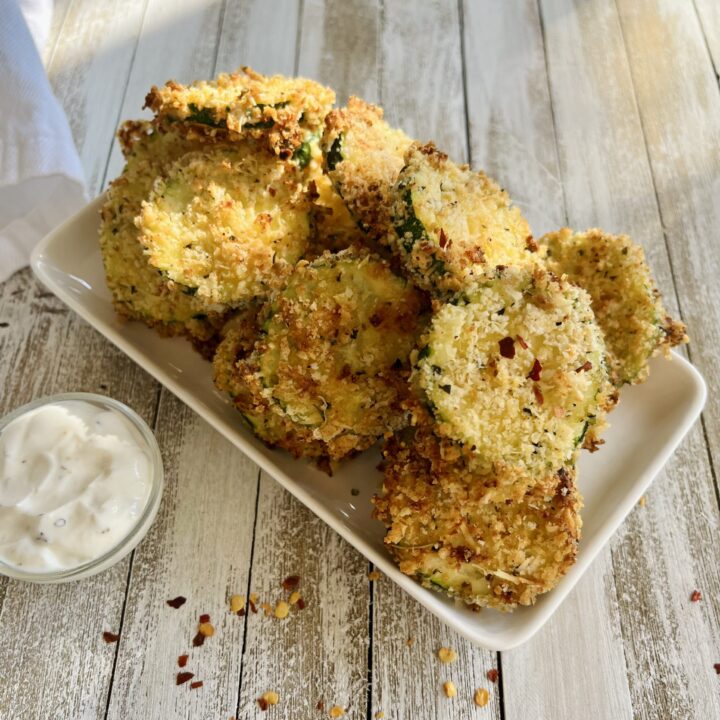 There is a small white rectangular dish with slices of baked zucchini slices on it. There is a very small bowl with a white creamy dressing in it. Items are on a wooden surface.