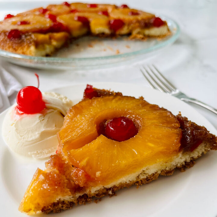 There is an upside down pineapple cake on a clear glass serving platter with a wedge missing from it. There is a small white plate with a piece of cake on it and a scoop of vanilla ice cream. There is a fork laying next to the plate. Items are on a white surface.