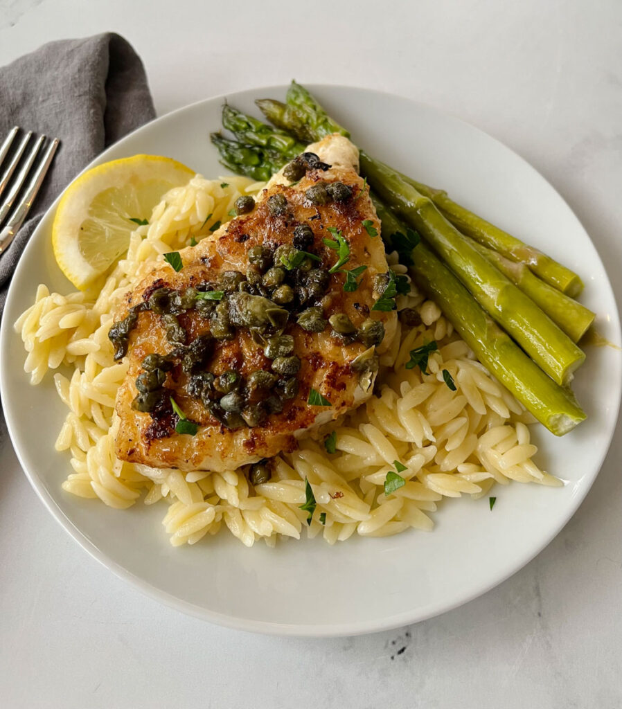There is a white plate with a bed of pasta, a piece of white fish with capers on it and stalks of asparagus. There is a gray cloth napkin laying next to the plate. Items are on a white surface.