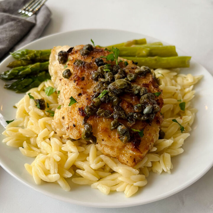 There is a white plate with a bed of pasta, a piece of white fish with capers on it and stalks of asparagus. There is a gray cloth napkin laying next to the plate. Items are on a white surface.