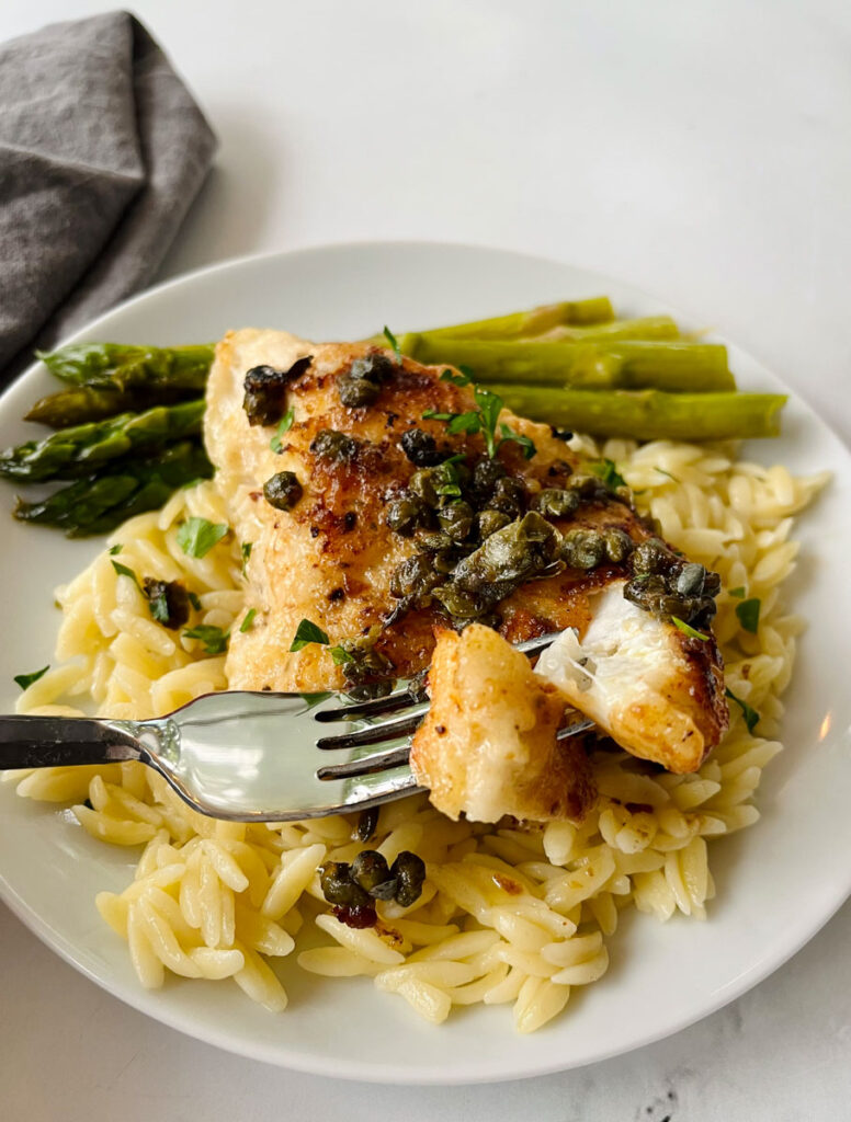 There is a white plate with a bed of pasta, a piece of white fish with capers on it and stalks of asparagus. There is a fork with a piece of the fish on it. There is a gray cloth napkin laying next to the plate. Items are on a white surface.