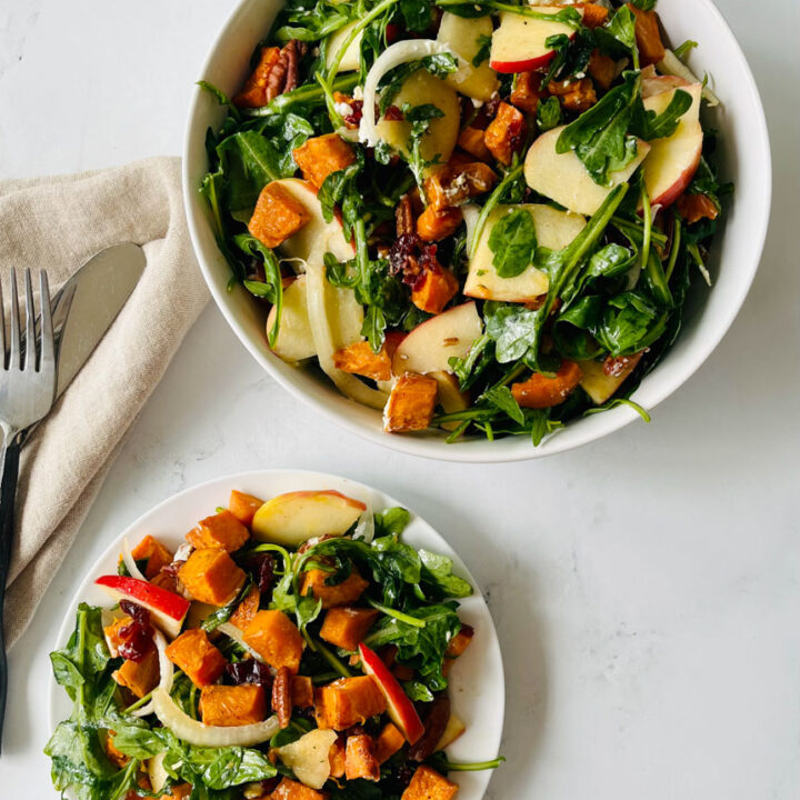 There is a small white plant and a large bowl with a salad mixture made up of arugula, roasted cubed sweet potatoes, goat cheese, sliced white onion, sliced apple, dried cranberries, and pecans tossed in a vinaigrette dressing. There is a tan cloth napkin, a fork, and knife laying next to the plate. Items are on a white surface.