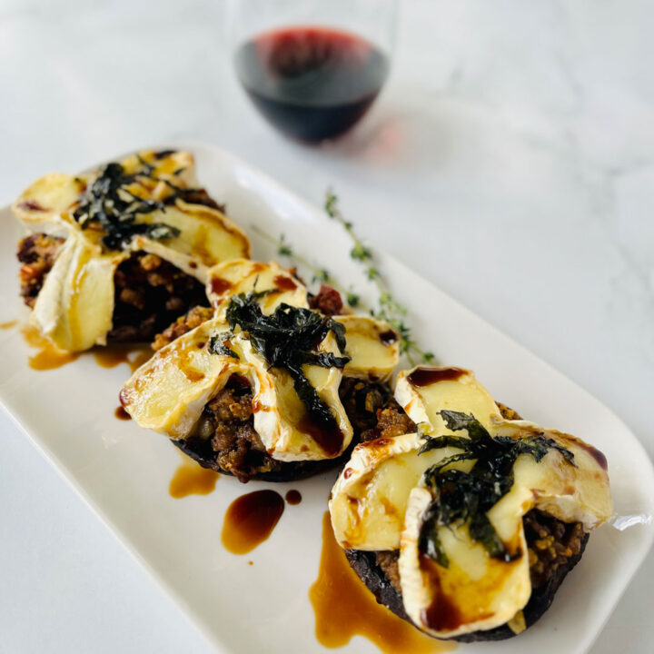 There is a large white rectangular plate with three large baked mushroom caps with stuffed with a sausage and cheese mixture. Caps have sliced cheese on and chopped basil on the top of them with balsamic vinegar drizzled over the plate. There is a glass of red wine in the background. Items are on a white surface.