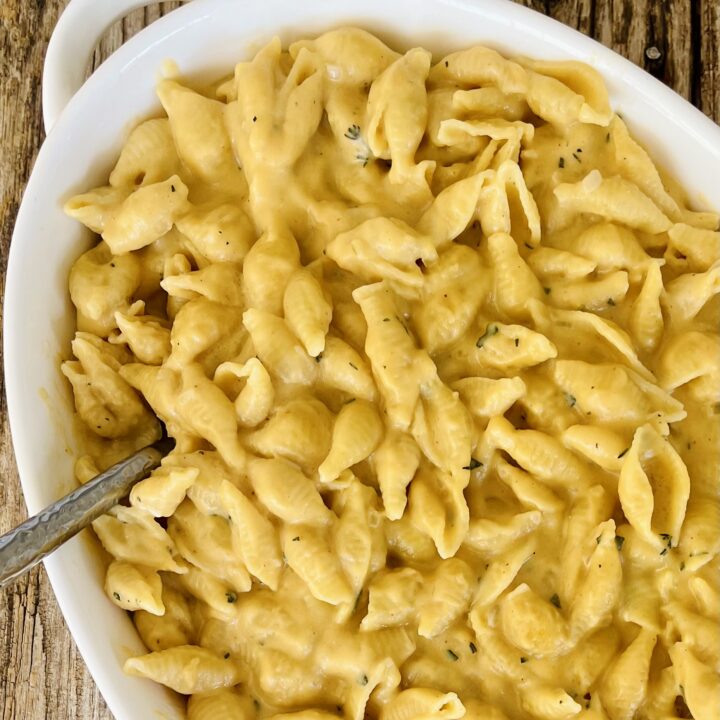 There is a large oval serving dish with Butternut Squash Mac and Cheese in it. There is a spoon resting in the dish. Dish is on a wood surface.