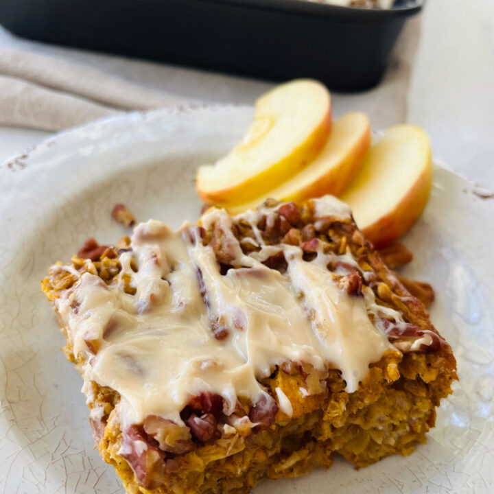 There is a piece of baked pumpkin-apple oatmeal with frosting on a plate. There are slices of apple on the plate. There is a black baking pan with the baked oatmeal in the background. Items are on a white surface.