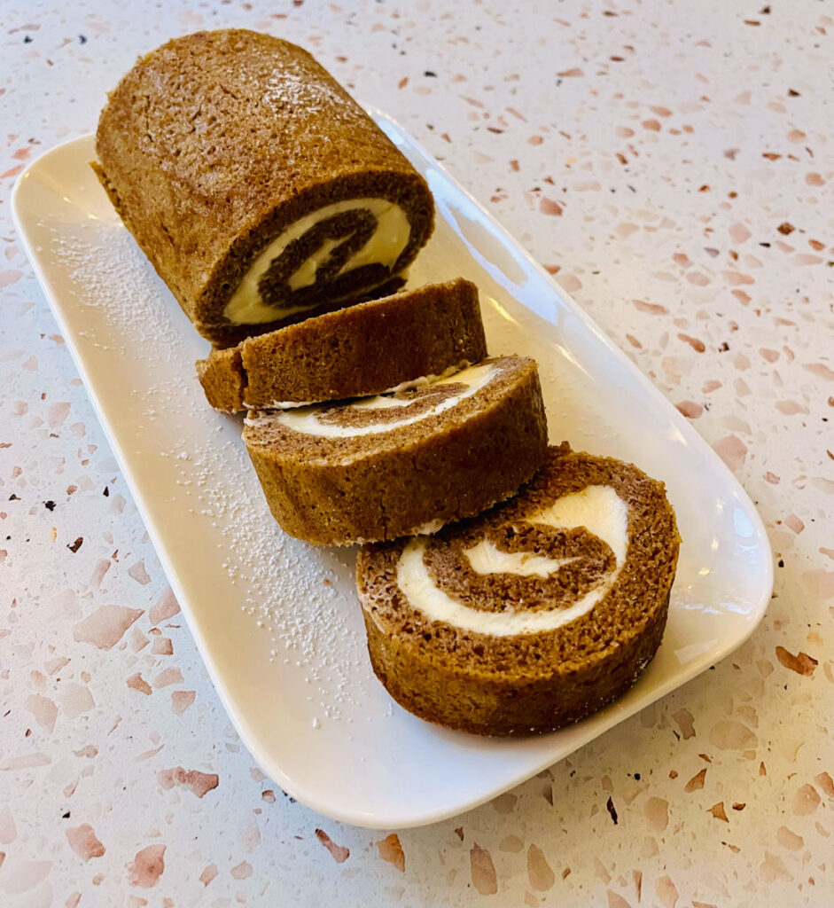 There is a small white rectangular platter with slices of a pumpkin roll on it. Platter is on an orange terrazzo surface.