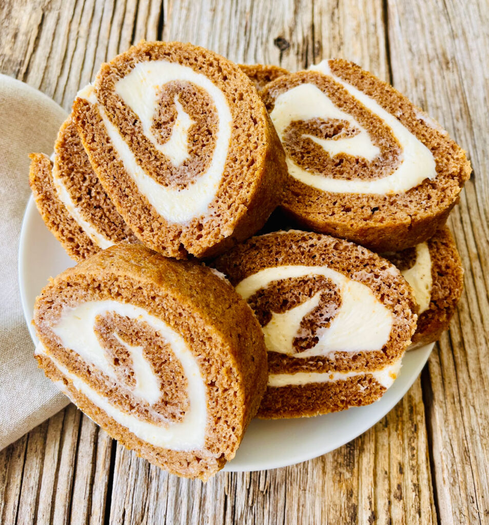 There is a white plate with slices of pumpkin roll piled on it. There is a tan cloth napkin next to the plate. Items are on a wood surface.