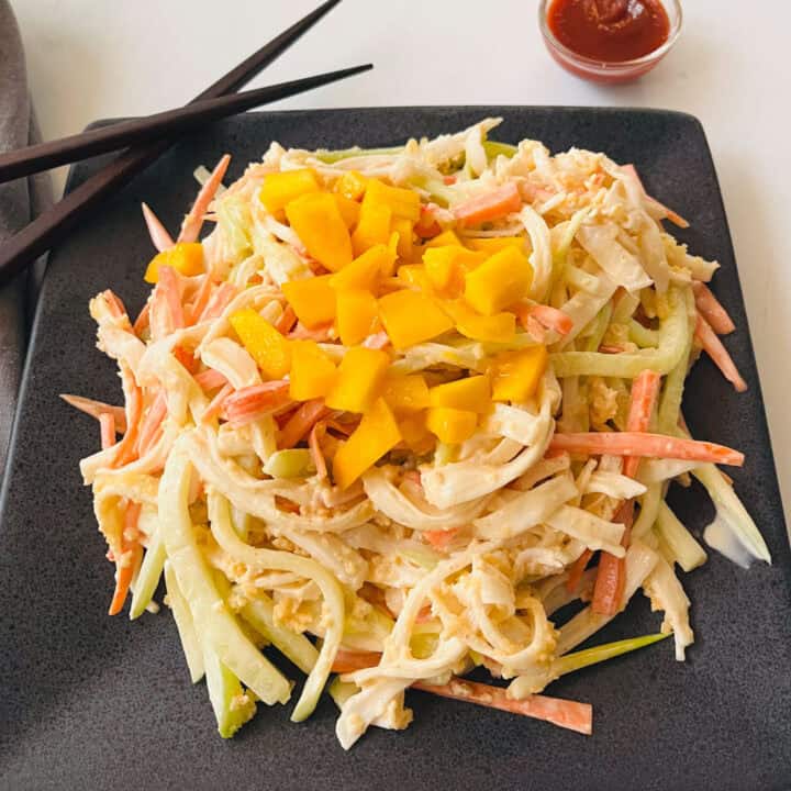 There is a black plate with a mound of kani salad: shredded imitation crab, cucumber, green onions, and carrots mixed in a dressing. There are chunks of fresh mango on top of the salad. There is a pair of chop sticks resting on the plate. There is a small bowl of red hot sauce in the background. Items are on a white surface.