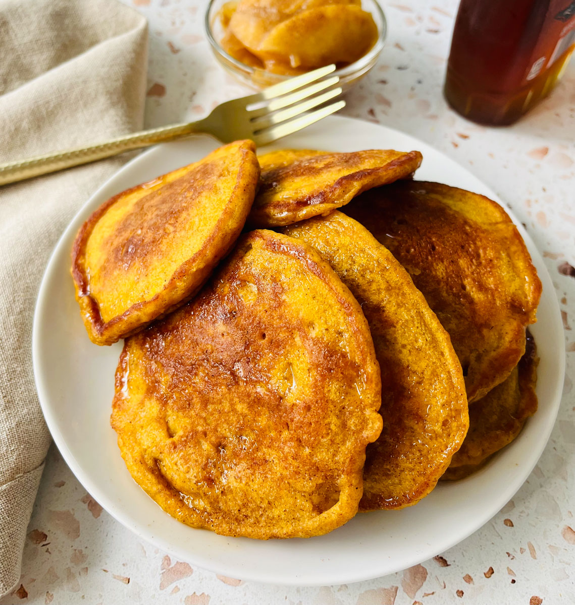 There is a white plate with a pile of pumpkin pancakes. There is a gold fork resting on the plate. There is a tan cloth napkin laying next to the plate. There is a small bowl of baked apples and a bottle of syrup in the background. Items are on an orange terrazzo surface.