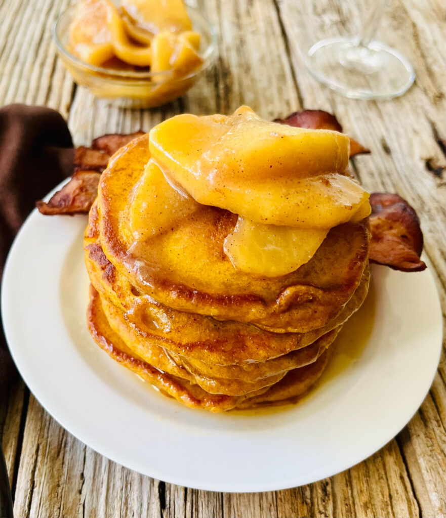 There is a white plate with a stack of pumpkin pancakes with a few baked apples and syrup on top of them. There are a few strips of bacon resting on the plate. There is a dark brown cloth napkin laying next to the plate. There is a small dish of baked apples in the background. Items are on a wood surface.