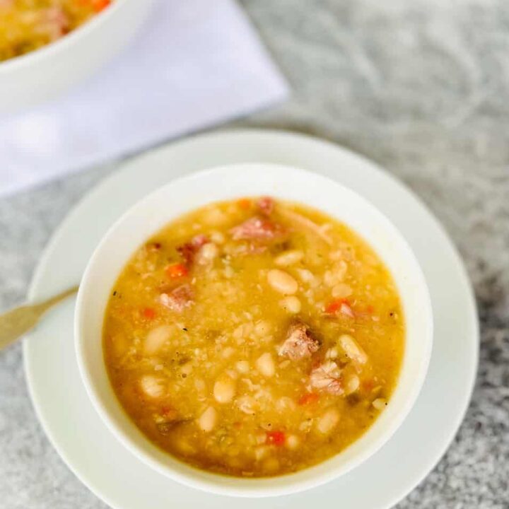 Small white round bowl with bean soup in it. Bowl is on a white round plate. There is a silver spoon resting on the plate. There is a large white round bowl with bean soup in it in the background. Bowls are on a light gray surface.