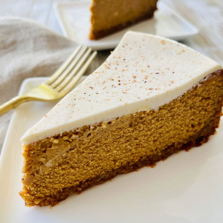 There is a wedge of pumpkin cheesecake with a sour cream topping on a white square plate. There is a gold fork resting on the plate. There is a tan cloth napkin laying next to the plate. In the background there is another white square plate with a wedge of cheesecake on it. Items are on a wood surface.