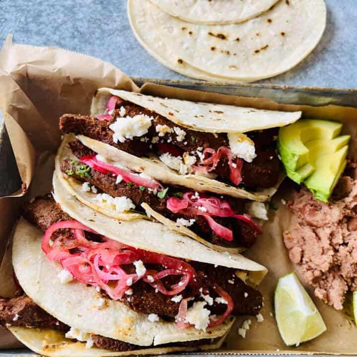There are three steak tacos with red onion and crumbled cheese, refried beans, slices of avocado, and a lime wedge on a paper-lined pan. There are corn tortillas laying in the background. Items are on a light gray surface.