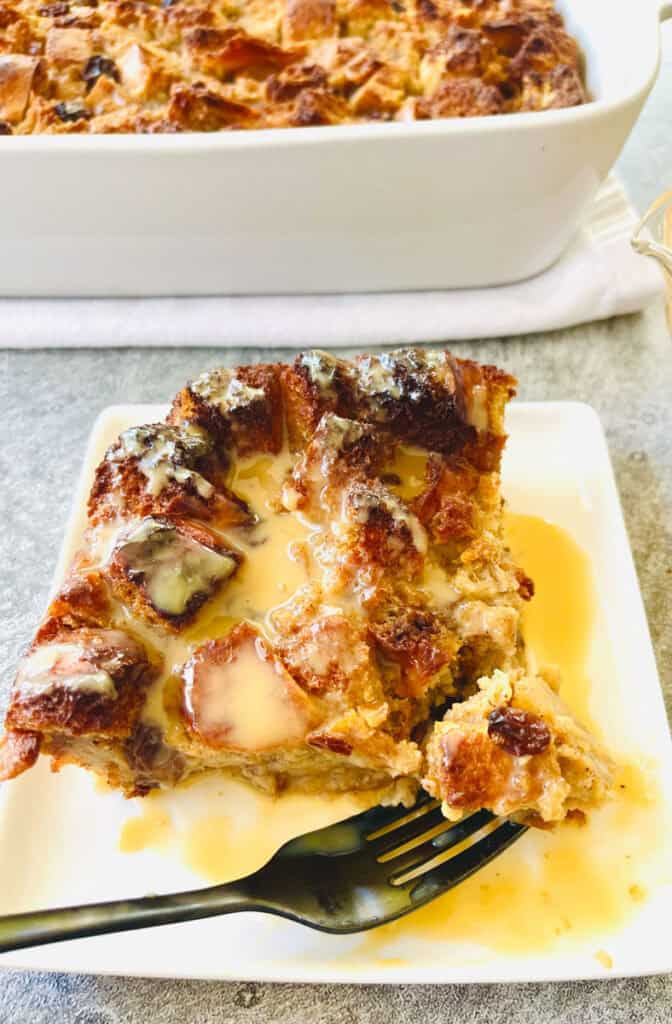 There is a square white plate with a piece of bread pudding with rum sauce on it. There is a fork resting on the plate. There is a white baking dish with bread pudding in it in the background. Items are on a light gray surface.