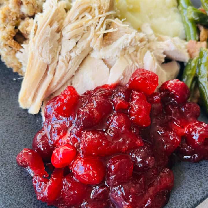 Black square plate with servings of cranberry sauce, sliced turkey, mashed potatoes, dressing, and green beans. There is a fork resting on the plate. Plate is on a wood surface.